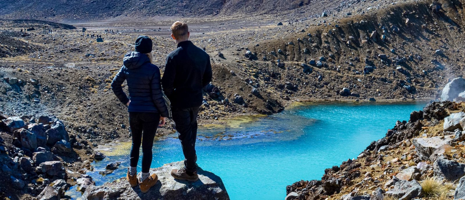Sabina and Liam standin on a rock in a volcanic landscape of Tongariro National Park looking out at the blue emerald lake, North Island, New Zealand