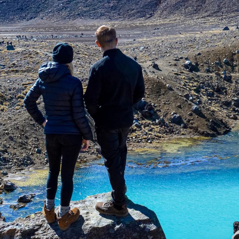 Sabina and Liam standin on a rock in a volcanic landscape of Tongariro National Park looking out at the blue emerald lake, North Island, New Zealand
