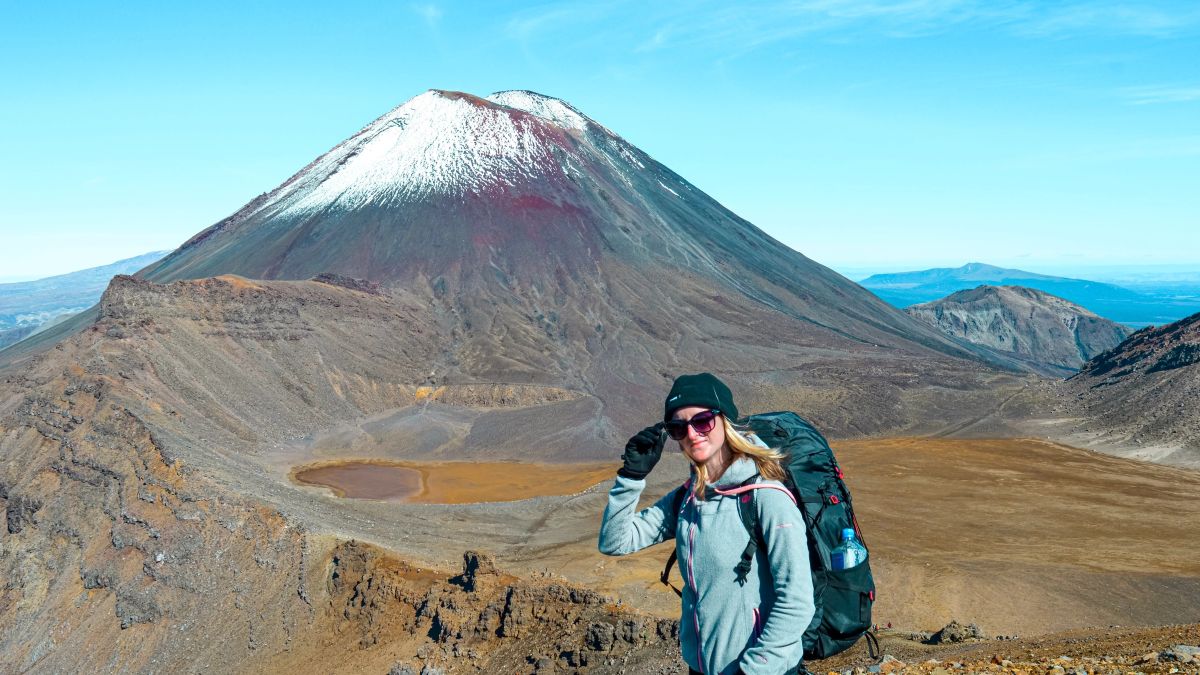 Sabina in full hiking gear standing on the age of volcanic landscape in front of a conical volcano called Mount Ngauruhoe (Mount Doom for LORT fans), Tongariro National Park, North Island, New Zealand