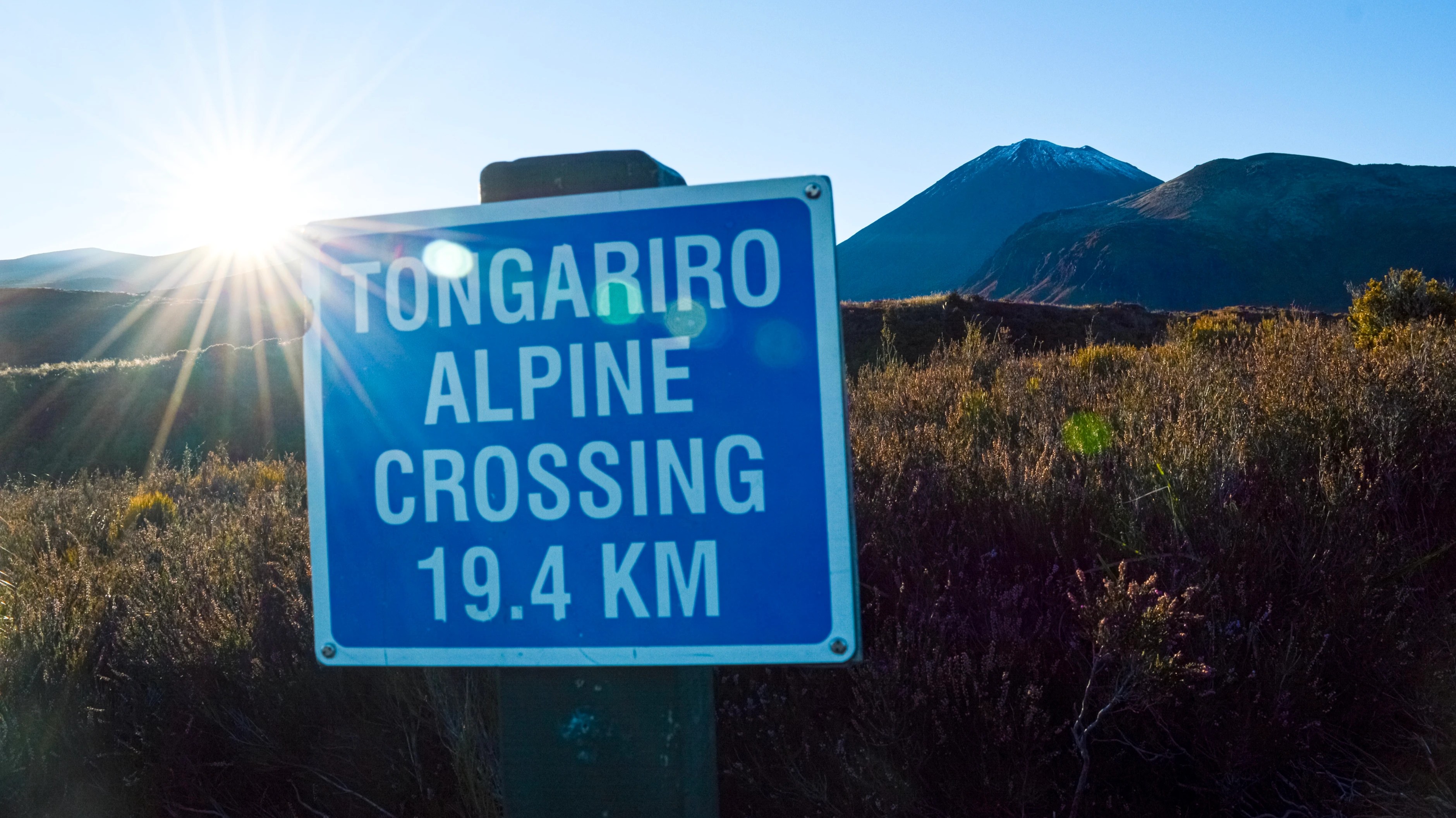 Tongariro Alpine Crossing start sign marking the 19.4 km hike illuminated by soft morning light with Mount Ngauruhoe on a clear day in New Zealand.