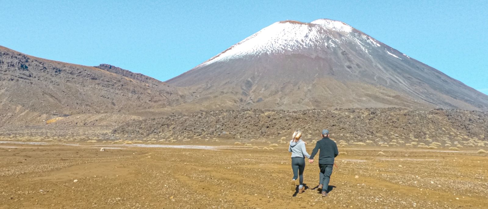 Sabina and Liam holding hands and running across an open plain with Mount Ngauruhoe in the background, North Island, New Zealand.