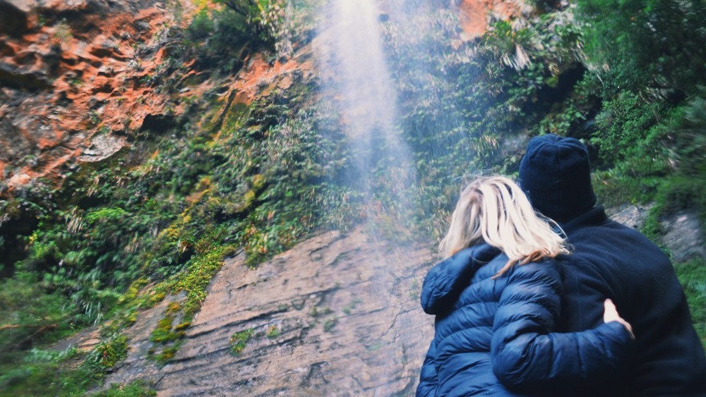 Couple standing at the base of Tupapakurua Falls watching the water falling down the rugged red rocks covered in green moss, North Island, New Zealand.
