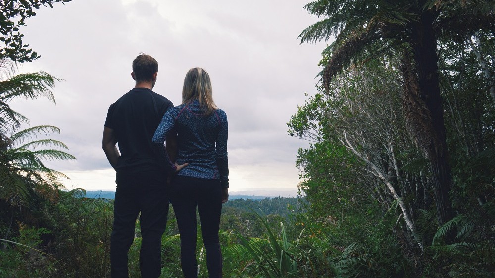 Sabina and Liam standing at the highest point of Tupapakurua Trail overlooking the vast green forest canopies, North Island, New Zealand.